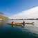 2 people in kayaks paddling through calm waters toward a mountainous coastal village